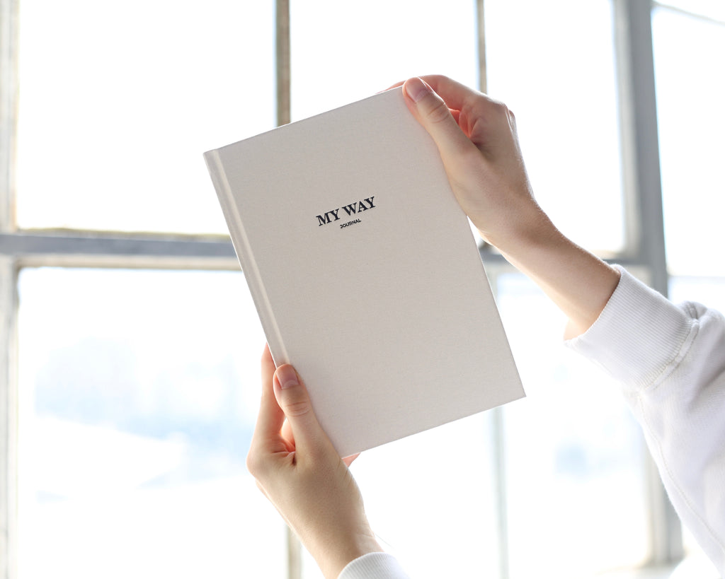 Hands holding the beige linen My Way Journal in front of a bright window, highlighting the premium textured cover of the My Way Journal undated planner and reflection journal.