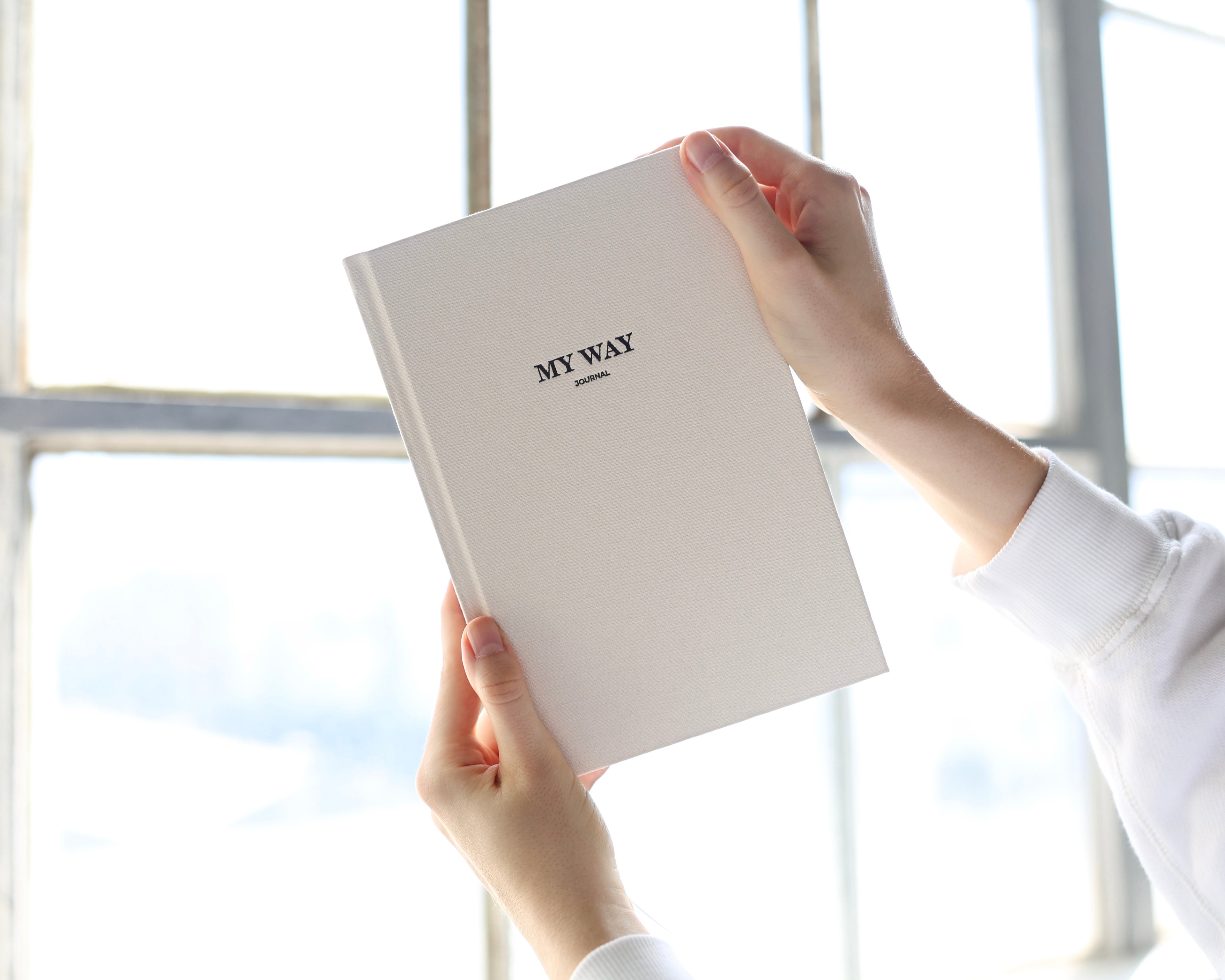 Hands holding the beige linen My Way Journal in front of a bright window, highlighting the premium textured cover of the My Way Journal undated planner and reflection journal.