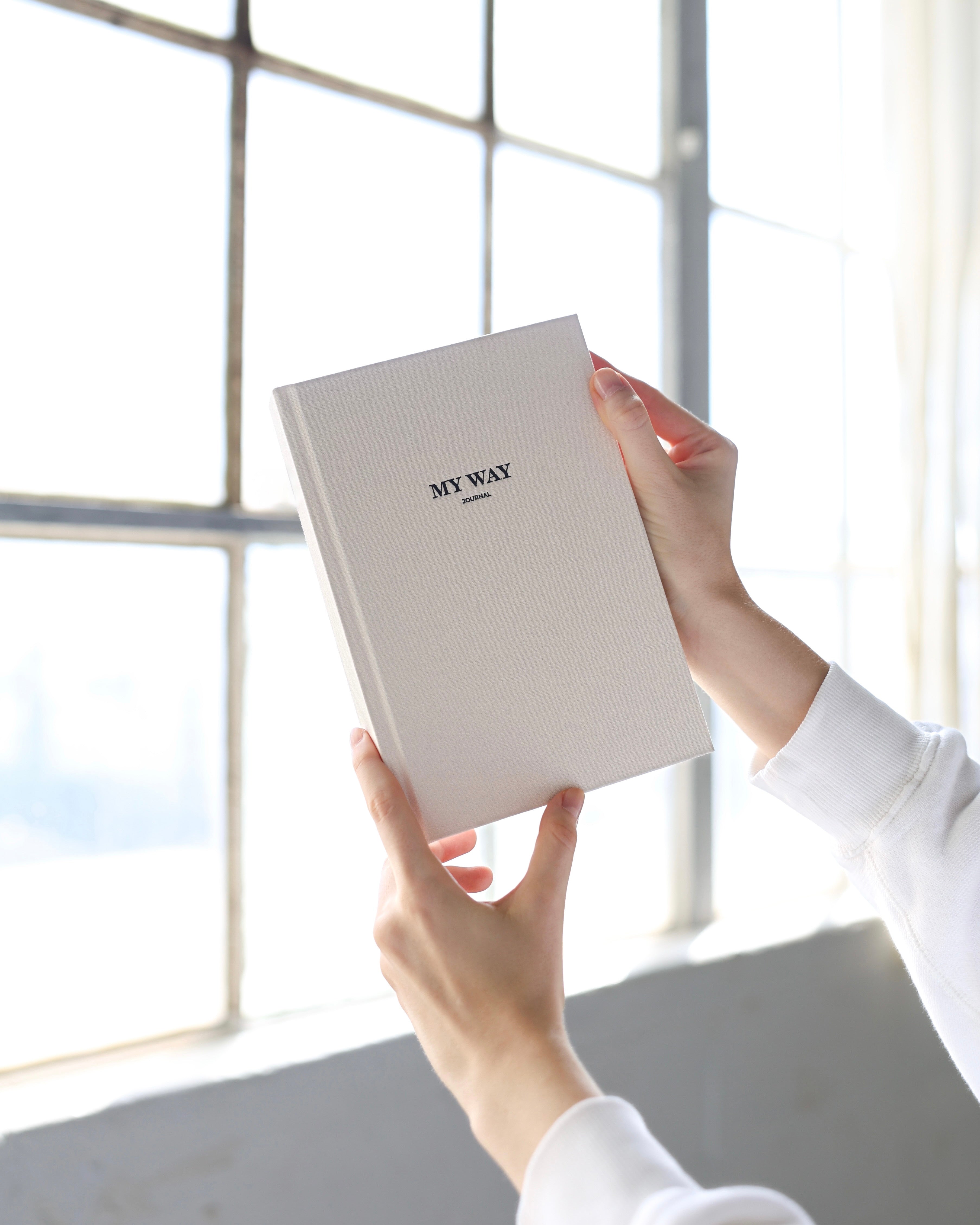 Hands holding the beige linen My Way Journal near a bright window, showing the premium textured cover and high-quality materials of the undated planner and reflection journal.
