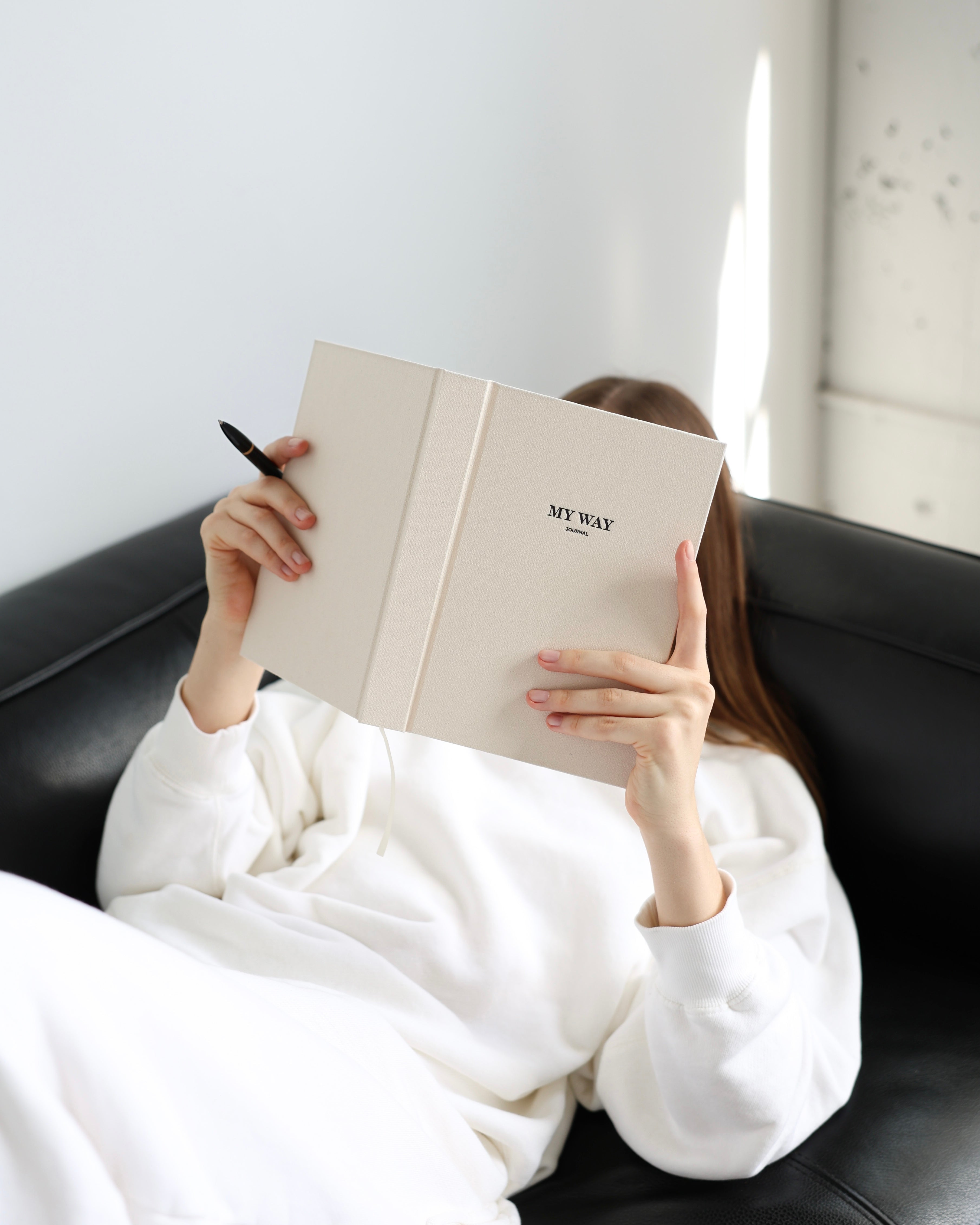 Person relaxing on a black sofa while holding a beige linen My Way Journal, minimalist lifestyle scene with premium undated planner and reflection journal.